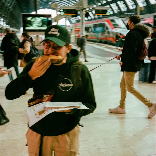 A man in a Jholano's Deli hoodie eats a sandwich at a busy train station.