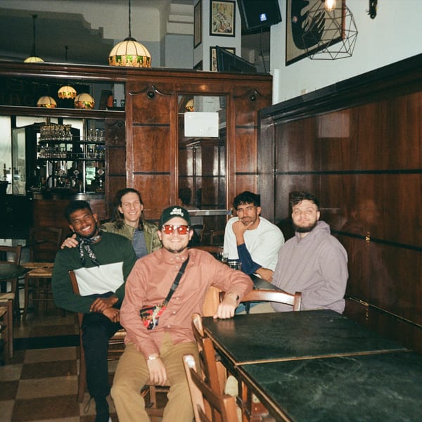Five young men smiling and posing together inside Jholano's Deli, with dark wood paneling and checkered floors.