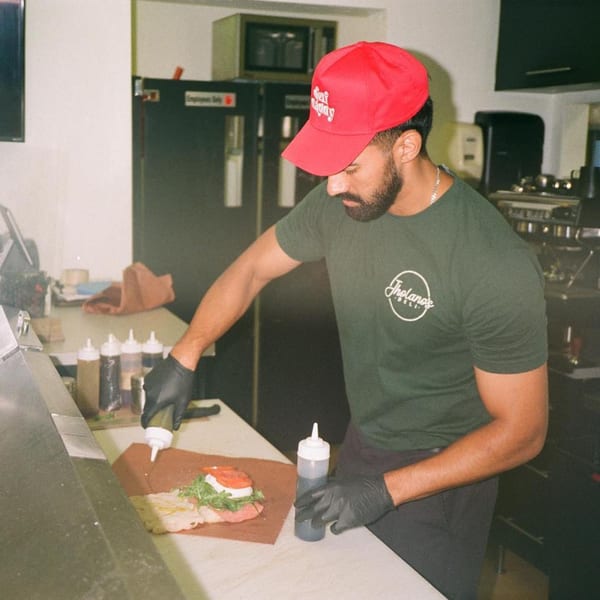 A Jholano's Deli employee in a red hat and green shirt prepares a sandwich with fresh ingredients.