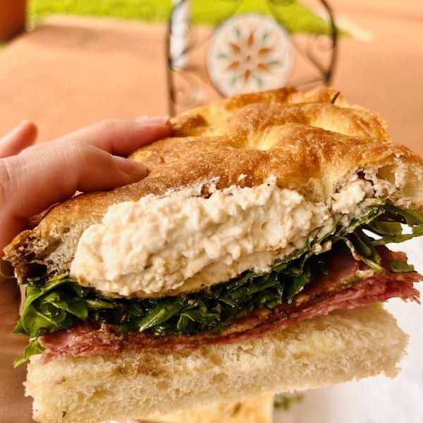 A hand holding a Jholano's Deli sandwich with focaccia, salami, arugula, and ricotta cheese.