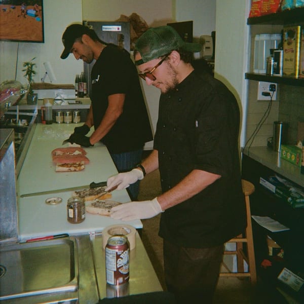 Two Jholano's Deli employees preparing sandwiches behind a counter in a deli kitchen.