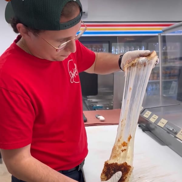 A Jholano's Deli employee in a red shirt stretches a grilled cheese sandwich, showing long, melted cheese pulls.