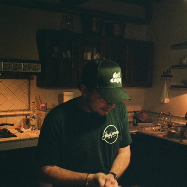 A person wearing a Jholano's Deli shirt and cap, preparing food in a dimly lit kitchen.