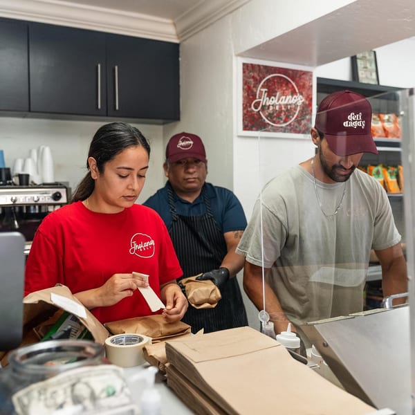 Three Jholano's Deli employees preparing sandwich orders behind a counter.