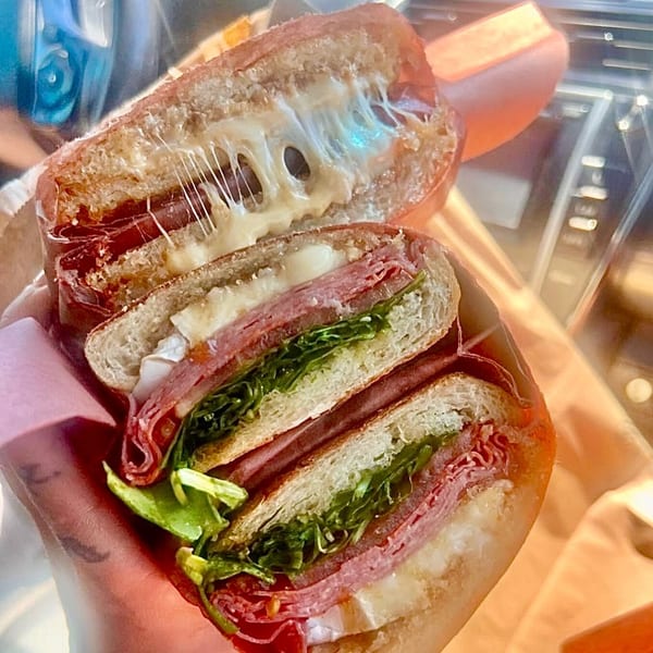 A Jholano's Deli sandwich, cut in half, revealing melted cheese, meats, and fresh greens.