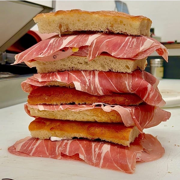A towering Jholano's Deli sandwich with focaccia bread, prosciutto, and mortadella on a white counter.