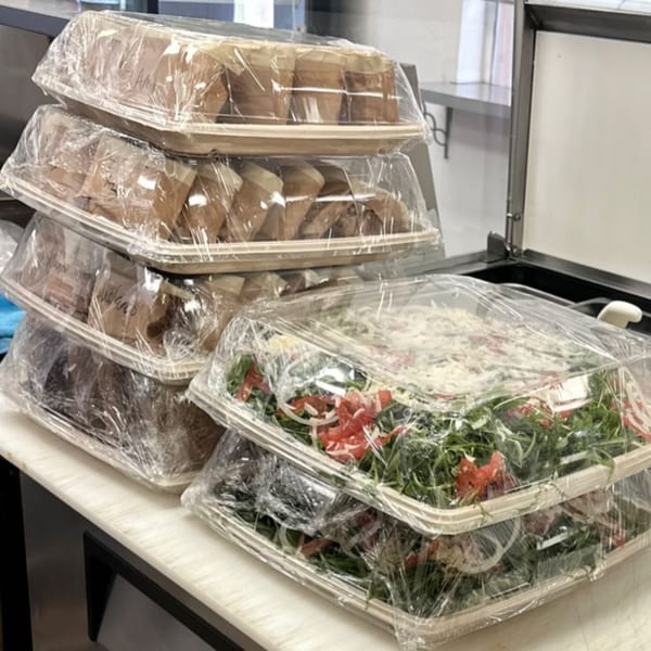 Catering order of Jholano's Deli sandwiches and salads in plastic-wrapped containers on a counter.