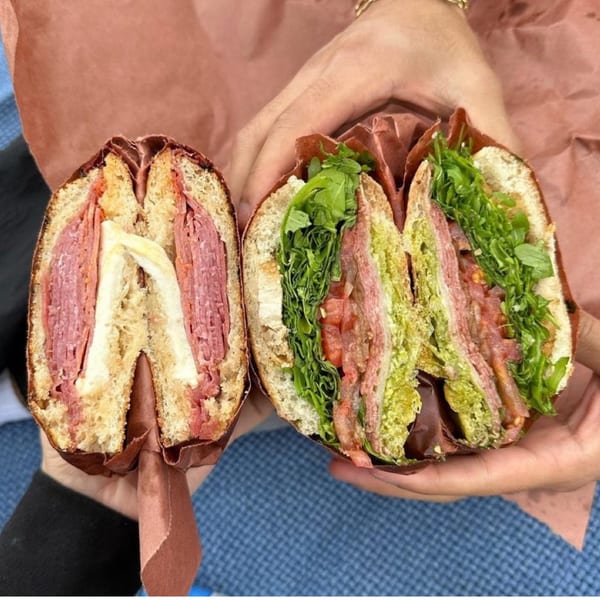 Two hands holding two halves of a Jholano's Deli sandwich, revealing layers of meat, cheese, and greens.