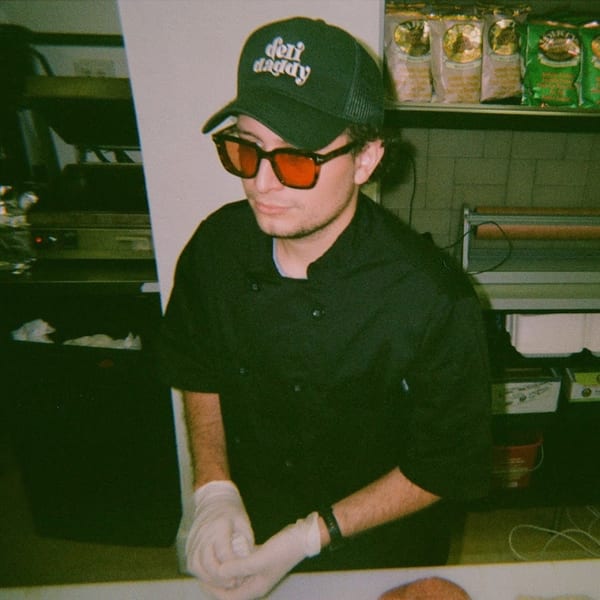 Jholano’s Deli employee wearing a "Deli Daddy" hat and red sunglasses, preparing food.